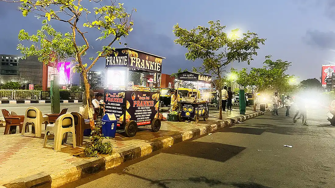 Frankie street food cart outside Surat VR Mall at dusk, with plastic chairs and auto rickshaws along the roadside