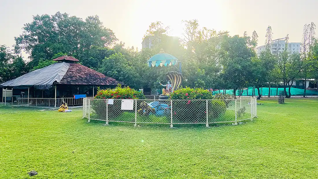 Green garden lawn at the ashram with a fenced central deity statue under a canopy, trees and a hut alongside
