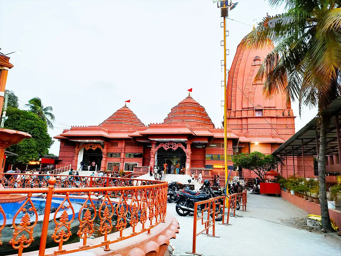 Red ISKCON Mandir with carved domes and saffron flags, fountain and parked bikes in the courtyard