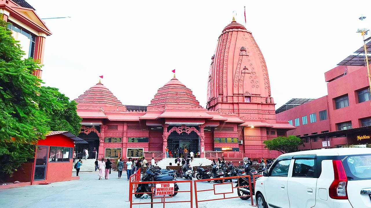 ISKCON Temple Surat with tall red domes and flags, parked vehicles and visitors near the two wheeler parking