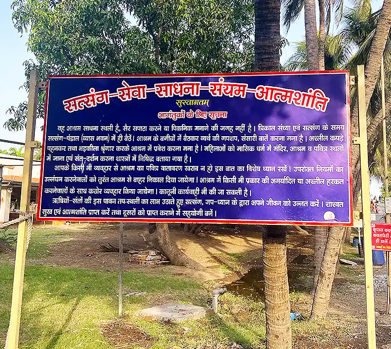 Blue information board at the ashram listing visitor guidelines in Hindi, set on a grassy patch among trees