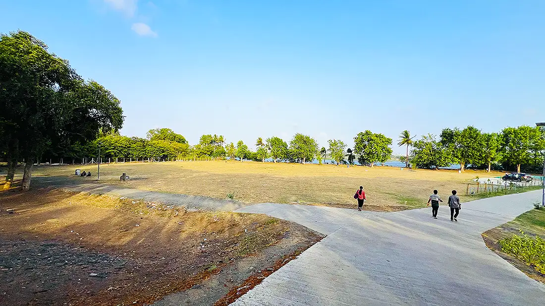 Wide open ground at the Jahangirpura ashram with a paved path, visitors walking and trees along the far edge