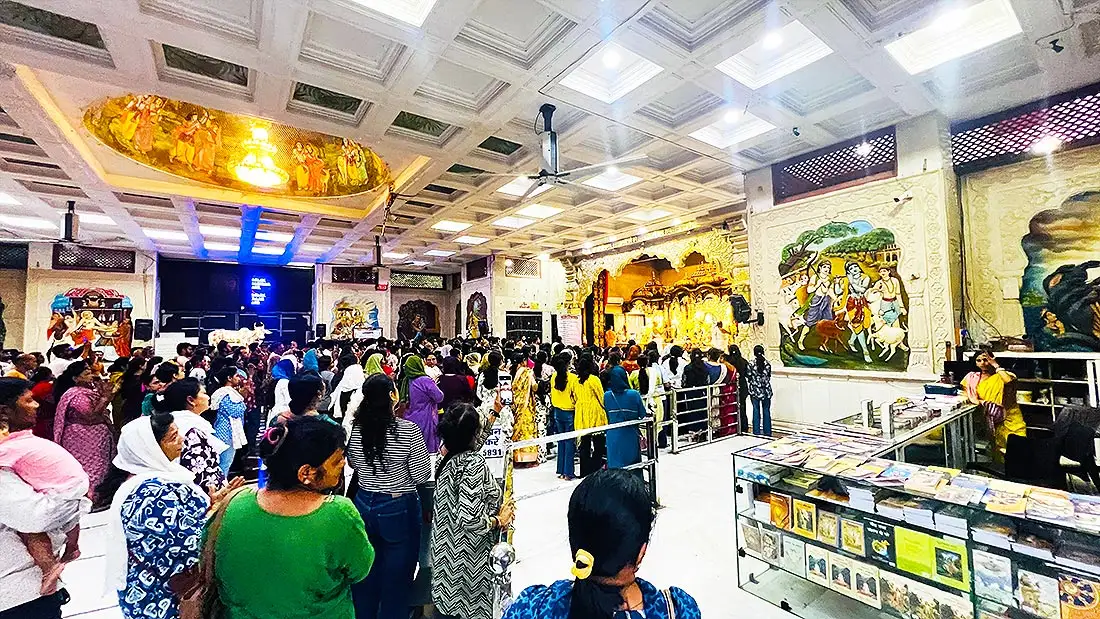 Devotees gathered for darshan inside ISKCON Temple hall with Krishna wall paintings and a book counter