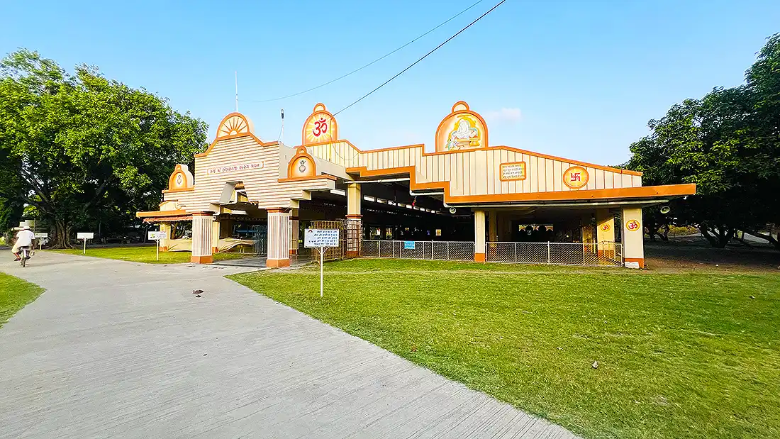 Side angle of the ashram satsang hall in Jahangirpura, cream and orange structure beside a wide green lawn and path