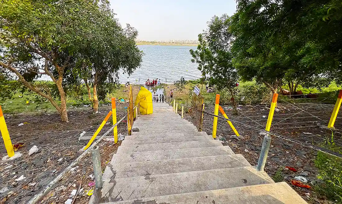 Stone steps leading down to the riverside at the Jahangirpura ashram, with people gathered near the water and trees around