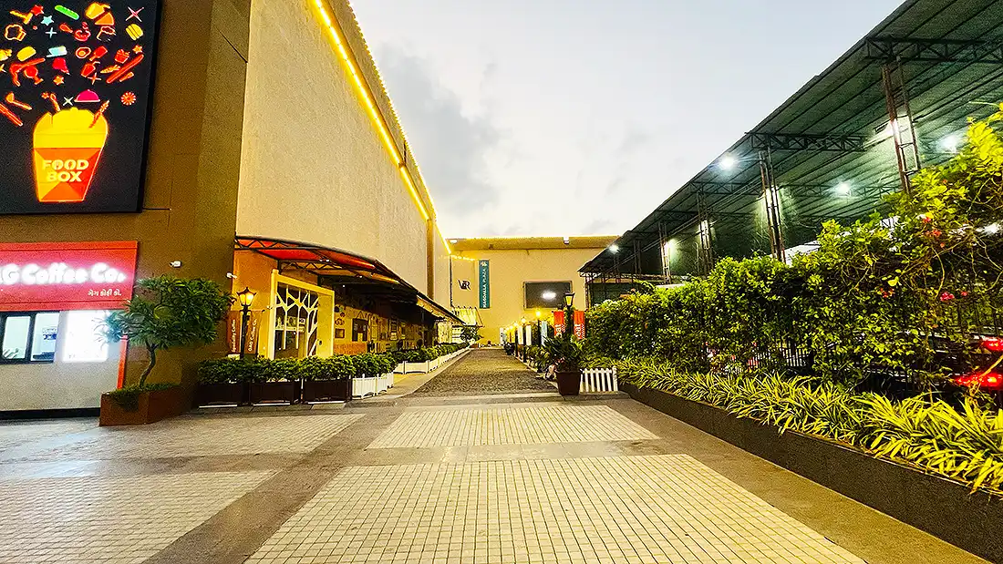 Side pathway at Surat VR Mall lit up in evening, lined with Food Box signage, plants and outdoor seating